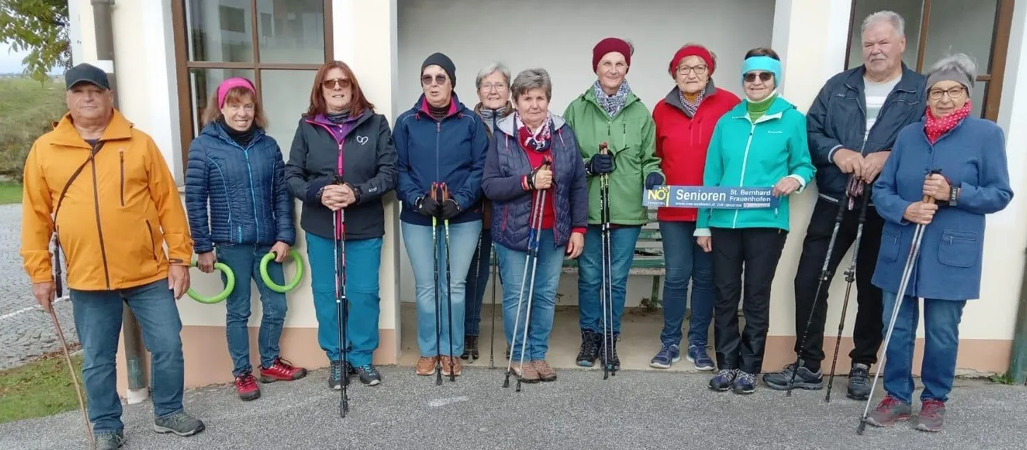 Eine Gruppe von Frauen, gekleidet in Outdoor-Ausrüstung, steht in einer Reihe, lächelt und hält Wanderstöcke. Hinter ihnen steht ein Gebäude mit einer Bank und einem Schild.