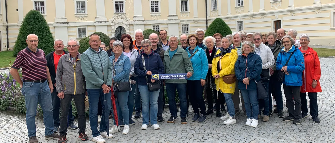 Eine Gruppe von Menschen steht vor einem großen Gebäude mit einem blauen Schild, auf dem NO! Senioren Horn steht. Sie alle lächeln und scheinen eine Tour zu machen.