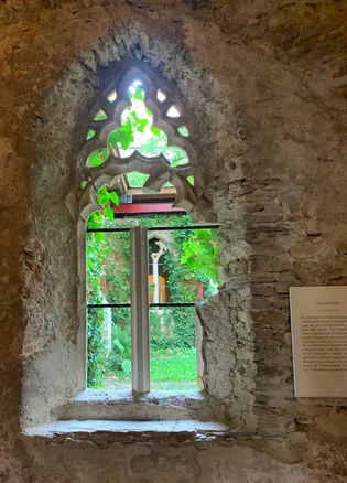 Eine steinerne Wand mit einem bogenförmigen Fenster und einem weißen Rahmen. Grüne Pflanzen bedecken das Fenster, und Sonnenlicht scheint hindurch. Ein Schild mit Text ist rechts angebracht.