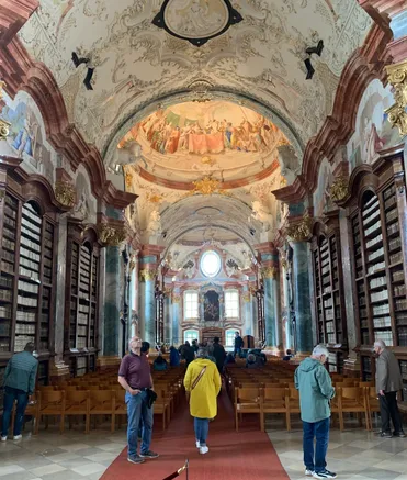 Eine prächtige Bibliothek mit gewölbten Decken und kunstvollen Gemälden. Menschen gehen zwischen den Reihen von Holzstühlen und Bücherregalen hindurch.