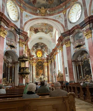 Das Innere einer großen Kirche mit kunstvollen Verzierungen, einschließlich eines goldenen runden Fensters und Fresken an der Decke. Eine Gruppe von Menschen sitzt auf den Kirchenbänken und schaut zum Altar.