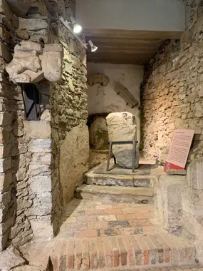 Ein kleiner, enger Raum mit Steinmauern und einem Ziegelboden, der eine große Steinplatte mit Inschriften zeigt. Steinartefakte sind an den Wänden montiert.