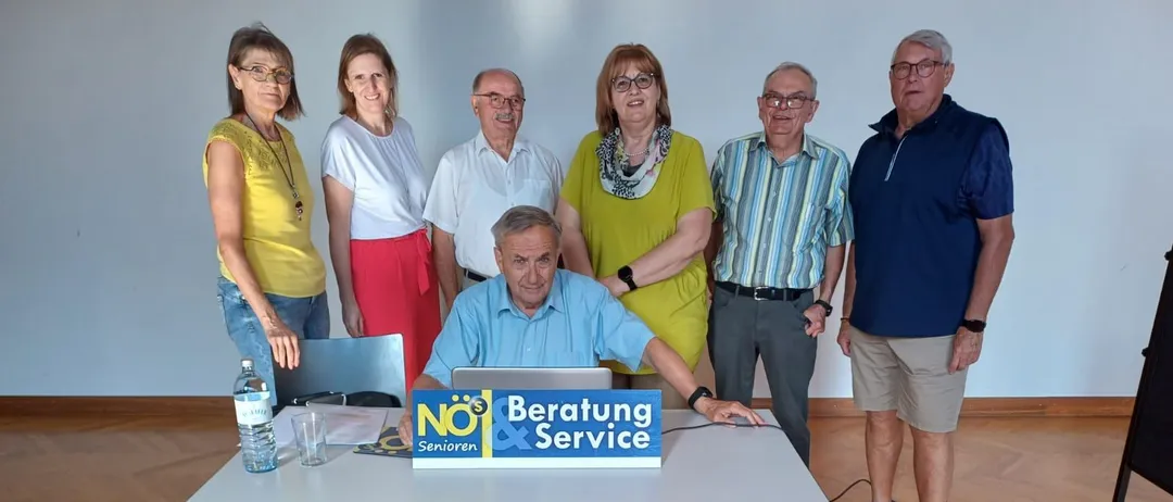 Eine Gruppe von Senioren versammelt sich um einen Tisch mit einem Laptop und posiert für ein Foto. Ein Schild mit NO$ Beratung & Service steht davor.