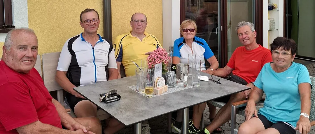 Sechs Menschen sitzen draußen an einem Tisch mit Getränken und Blumen. Zwei Frauen und vier Männer tragen Freizeitkleidung. Sie sitzen unter einem Vordach mit einer gelben Wand.