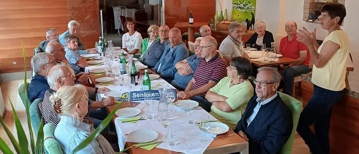 Eine Gruppe älterer Menschen sitzt um einen langen Esstisch in einem Esszimmer, einige unterhalten sich. Flaschen und Gläser stehen auf dem Tisch. Eine Frau steht in der Nähe und lächelt.