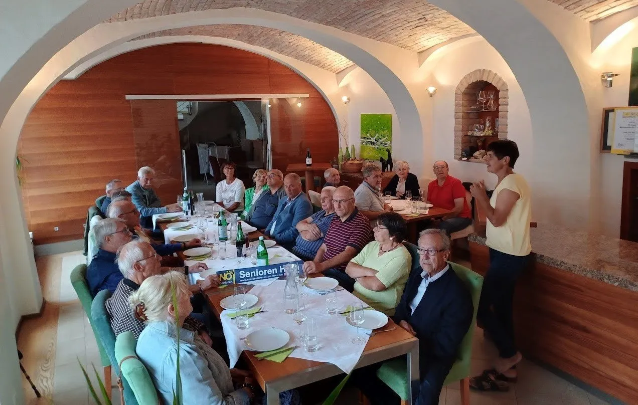 Eine Gruppe älterer Menschen sitzt um einen Tisch in einem Restaurant herum, möglicherweise auf einer Versammlung. Es gibt Flaschen, Gläser und Teller auf dem Tisch. Das Restaurant hat Holzwände, ein Backsteinregal und Lampen. Eine Frau steht vor ihnen.