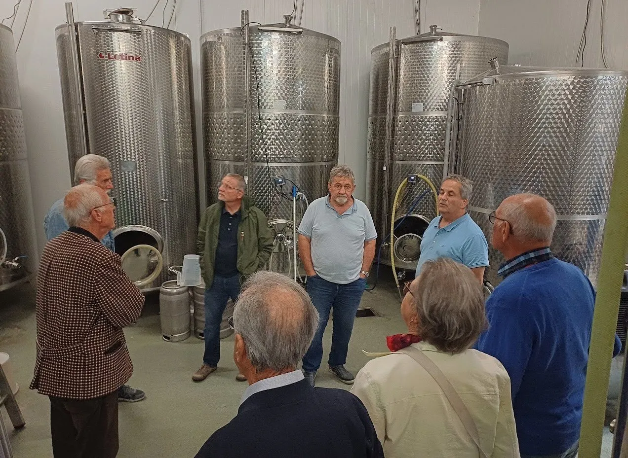 Eine Gruppe von Männern und Frauen versammelt sich in einem Raum mit großen Edelstahltanks. Sie tragen Freizeitkleidung und scheinen etwas zu besprechen. Der Raum hat eine weiße Wand.