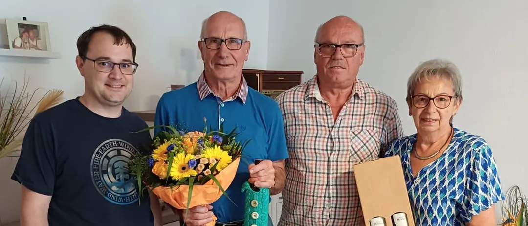 Vier Menschen stehen in einem Raum zusammen. Der Mann auf der linken Seite hält einen Blumenstrauß. Der Mann in der Mitte trägt ein blaues Polo-Shirt und hält ein grünes Band. Der Mann auf der rechten Seite trägt ein kariertes Hemd und hält eine Box. Die Frau auf der rechten Seite hält einen weißen Umschlag.