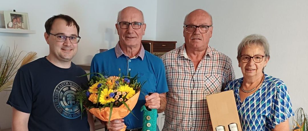 Vier Menschen stehen in einem Raum zusammen. Der Mann auf der linken Seite hält einen Blumenstrauß. Der Mann in der Mitte trägt ein blaues Polo-Shirt und hält ein grünes Band. Der Mann auf der rechten Seite trägt ein kariertes Hemd und hält eine Box. Die Frau auf der rechten Seite hält einen weißen Umschlag.