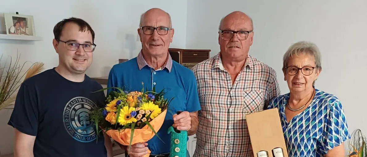 Vier Menschen stehen in einem Raum zusammen. Der Mann auf der linken Seite hält einen Blumenstrauß. Der Mann in der Mitte trägt ein blaues Polo-Shirt und hält ein grünes Band. Der Mann auf der rechten Seite trägt ein kariertes Hemd und hält eine Box. Die Frau auf der rechten Seite hält einen weißen Umschlag.