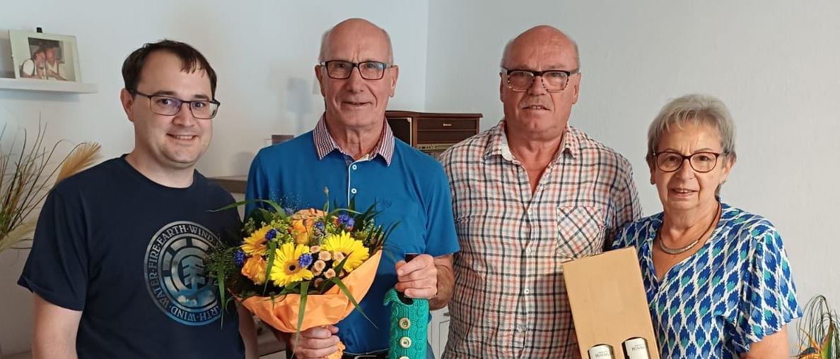Vier Menschen stehen in einem Raum zusammen. Der Mann auf der linken Seite hält einen Blumenstrauß. Der Mann in der Mitte trägt ein blaues Polo-Shirt und hält ein grünes Band. Der Mann auf der rechten Seite trägt ein kariertes Hemd und hält eine Box. Die Frau auf der rechten Seite hält einen weißen Umschlag.