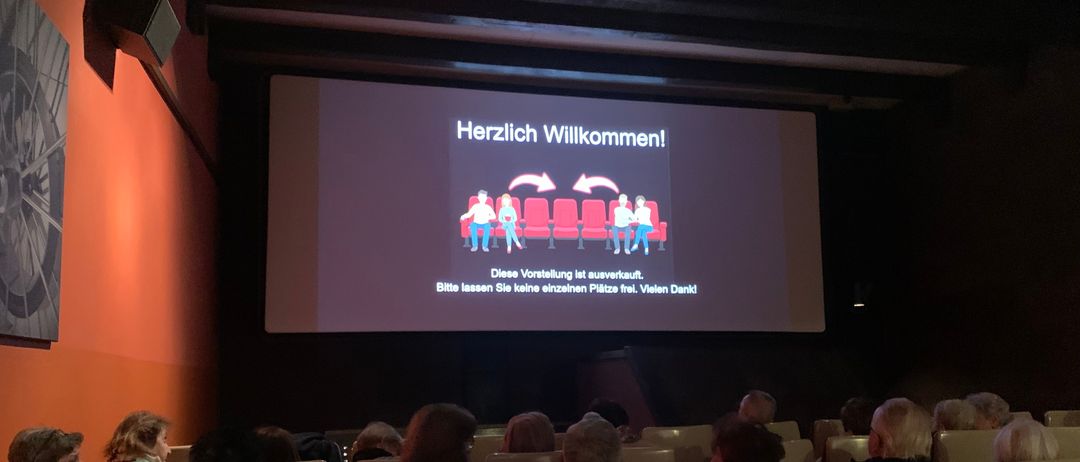 Ein Publikum sitzt in einem Kinosaal und schaut sich eine Präsentationsfolie auf einer Leinwand an, die 'Herzlich Willkommen!' mit einer Illustration von Menschen auf Stühlen sagt.