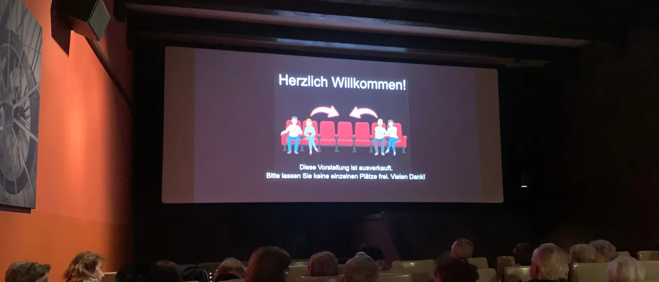 Ein Publikum sitzt in einem Kinosaal und schaut sich eine Präsentationsfolie auf einer Leinwand an, die 'Herzlich Willkommen!' mit einer Illustration von Menschen auf Stühlen sagt.