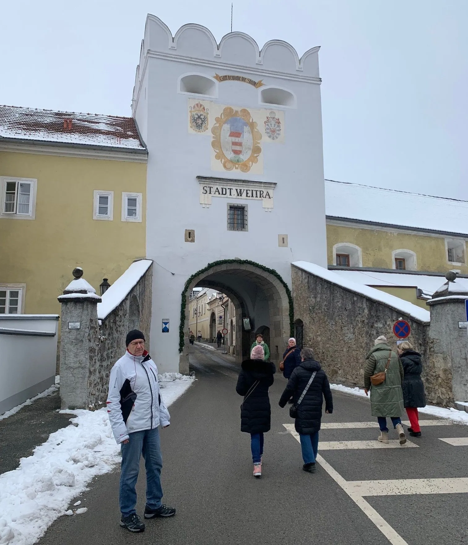 Eine Gruppe von Menschen geht vor dem Stadt Weitra Tor. Das Tor hat ein Wappen und den Text Stadt Weitra. Die Leute tragen Winterkleidung und haben Taschen.