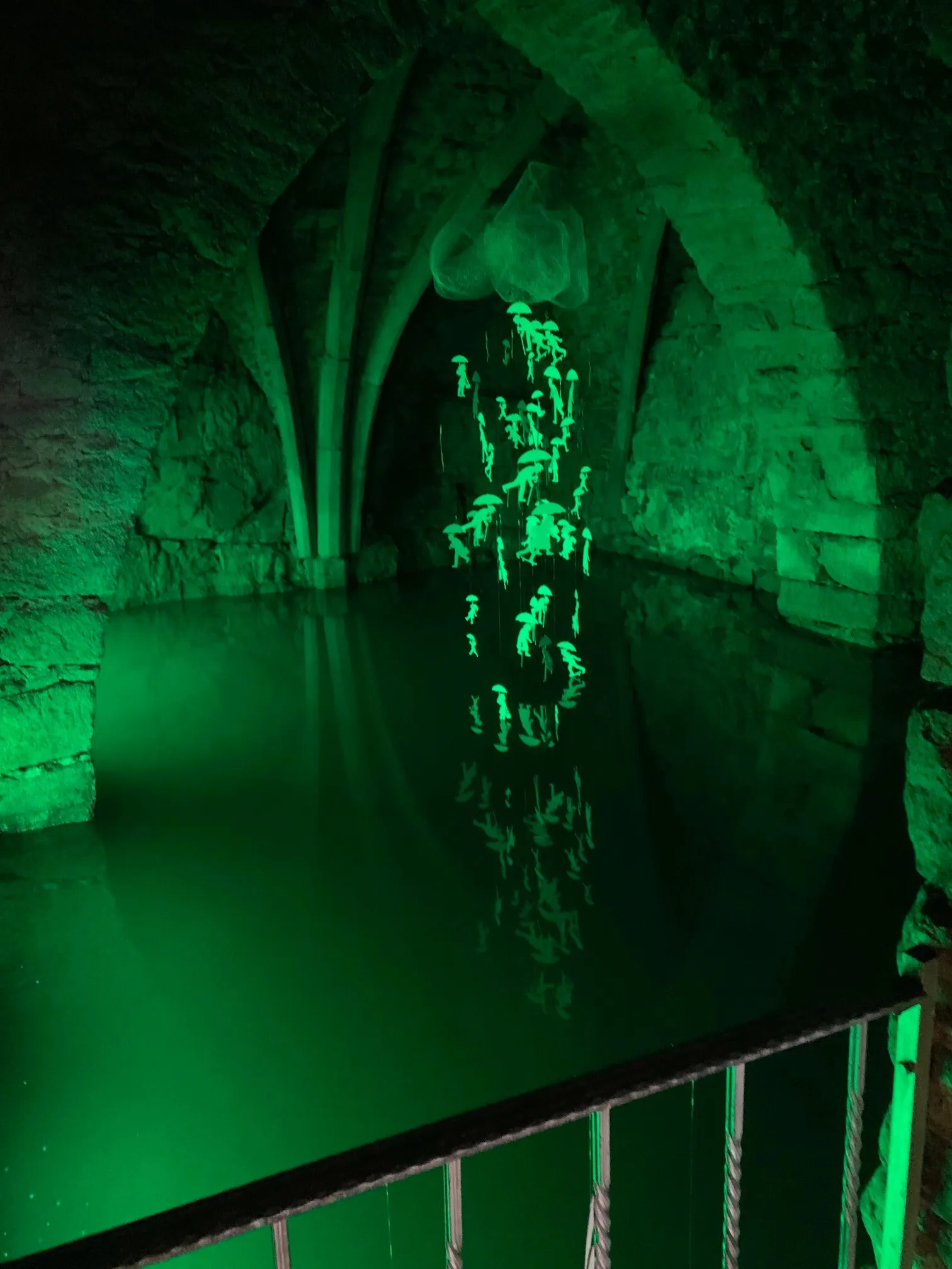 Ein schwach beleuchteter unterirdischer Pool mit grüner Beleuchtung hebt eine leuchtende Kronleuchter-artige Struktur im Wasser hervor. Steinwände umgeben den Pool.