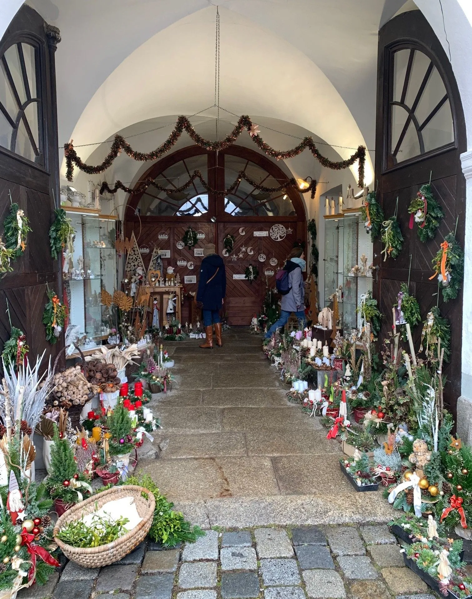 Zwei Personen in Winterkleidung erkunden einen festlichen Markt mit einer Vielzahl von Kränzen, Ornamenten und Weihnachtsdekorationen. Eine Holztür mit kunstvollen Schnitzereien und Lichtern verleiht dem Ganzen zusätzliches Flair.