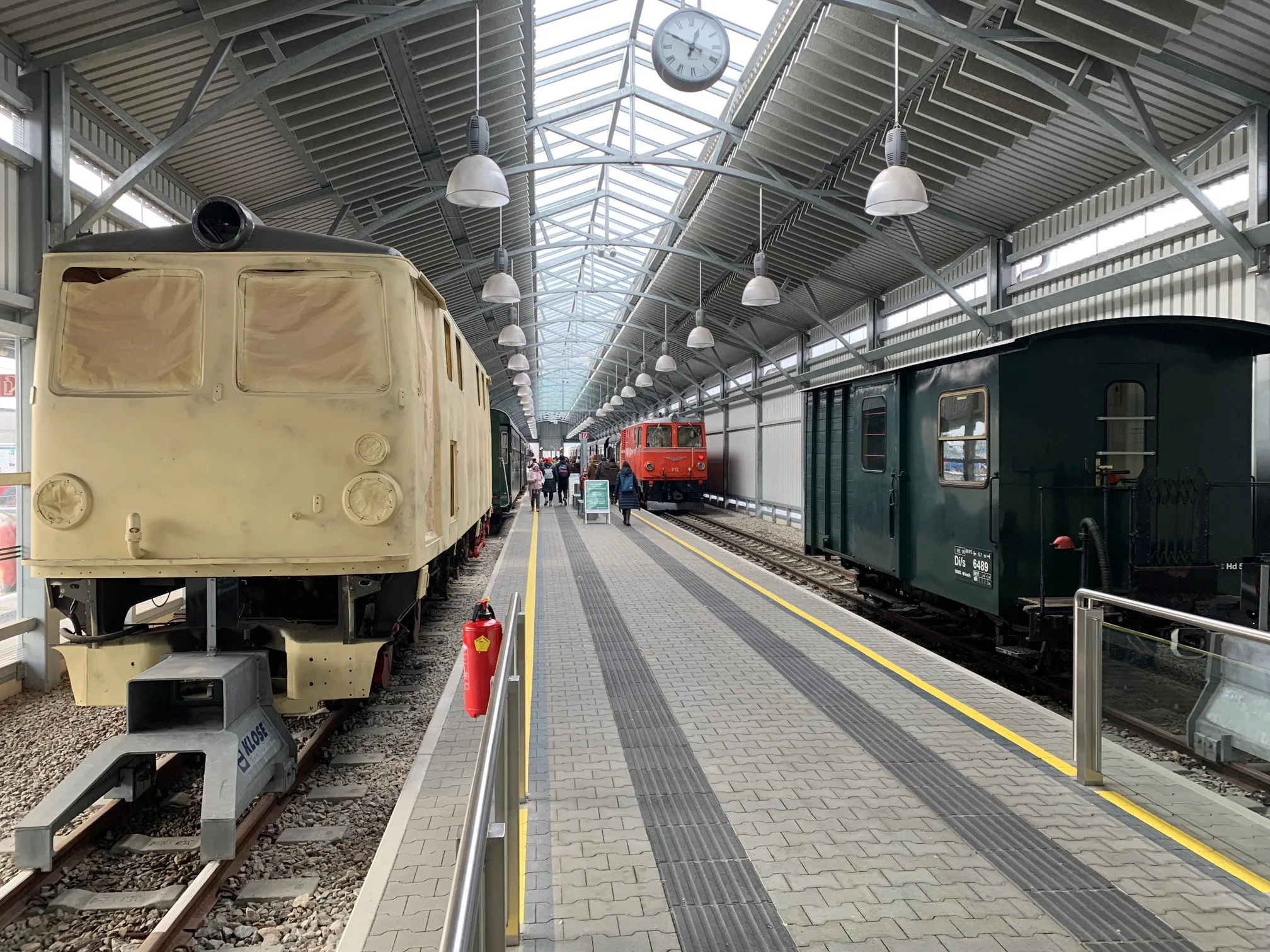 Ein überdachter Bahnhof mit mehreren geparkten Zügen. Der Bahnhof hat einen Ziegelboden und gelbe Linien. Es gibt hängende Lampen und eine Uhr darüber.