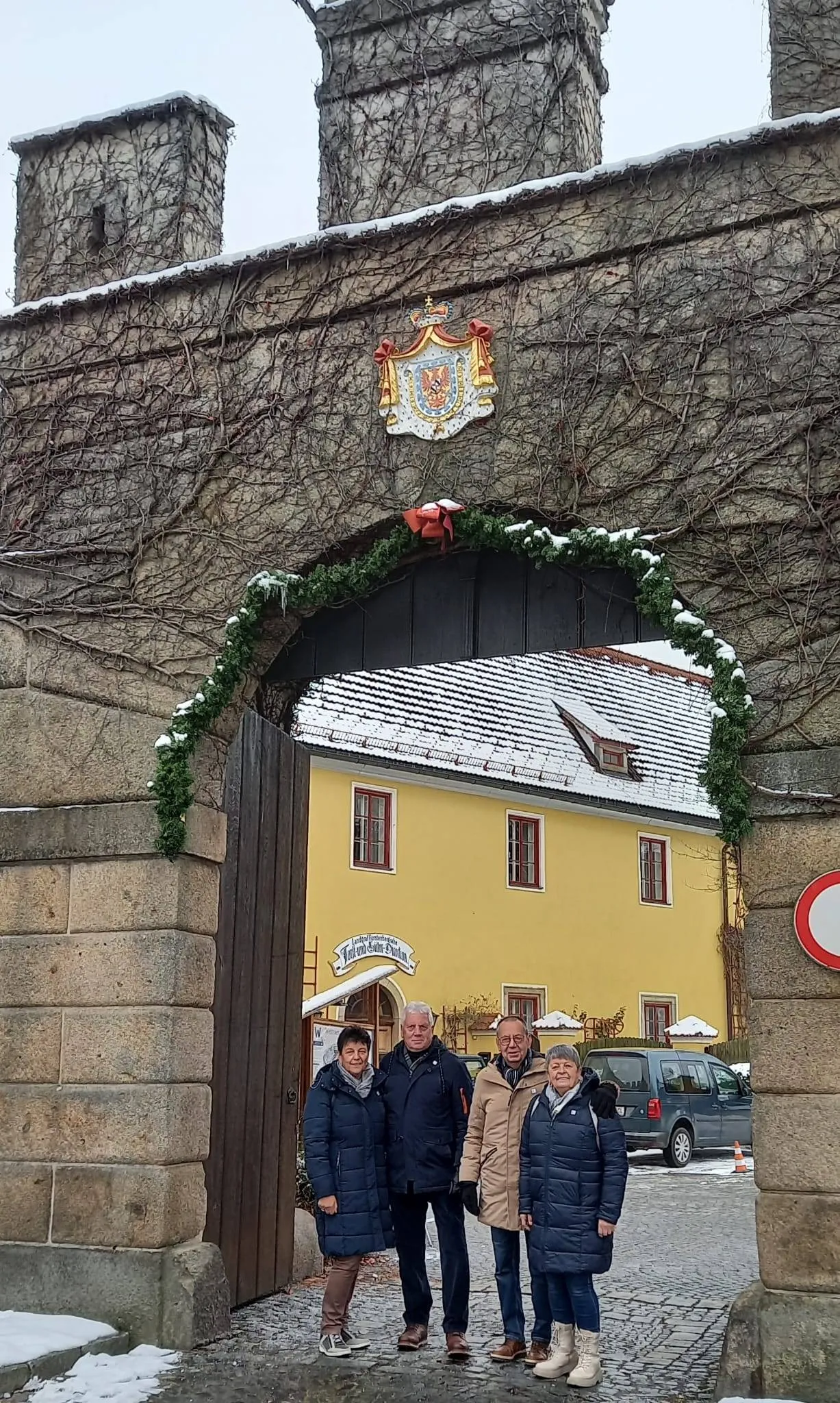 Drei Personen stehen vor einem gelben Gebäude mit Schnee auf dem Dach, unter einem Steinbogen mit Grün. Ein Wappenschild ist über dem Bogen.