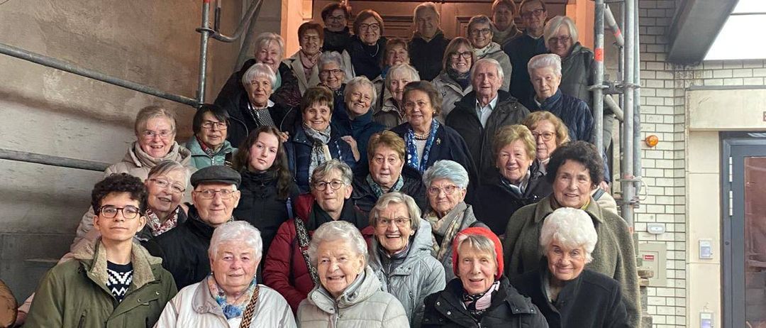 Eine Gruppe älterer Menschen, sowohl Männer als auch Frauen, stehen vor einem Gebäude mit Gerüst. Einige tragen Brillen, Mützen und Schals. Sie scheinen für ein Foto zu posieren.