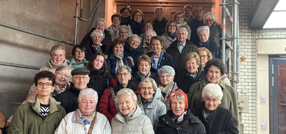 Eine Gruppe älterer Menschen, sowohl Männer als auch Frauen, stehen vor einem Gebäude mit Gerüst. Einige tragen Brillen, Mützen und Schals. Sie scheinen für ein Foto zu posieren.
