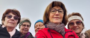 Fünf Frauen stehen lächelnd zusammen. Die Frau in der Mitte trägt eine Brille und einen Schal. Hinter ihr ist eine Frau mit einer blauen Mütze zu sehen. Der Himmel ist bedeckt.