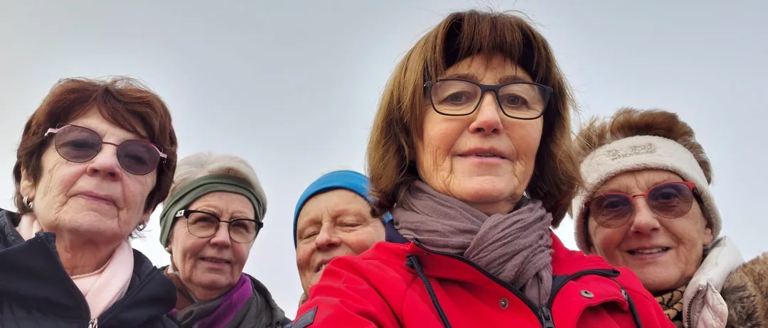Fünf Frauen stehen lächelnd zusammen. Die Frau in der Mitte trägt eine Brille und einen Schal. Hinter ihr ist eine Frau mit einer blauen Mütze zu sehen. Der Himmel ist bedeckt.