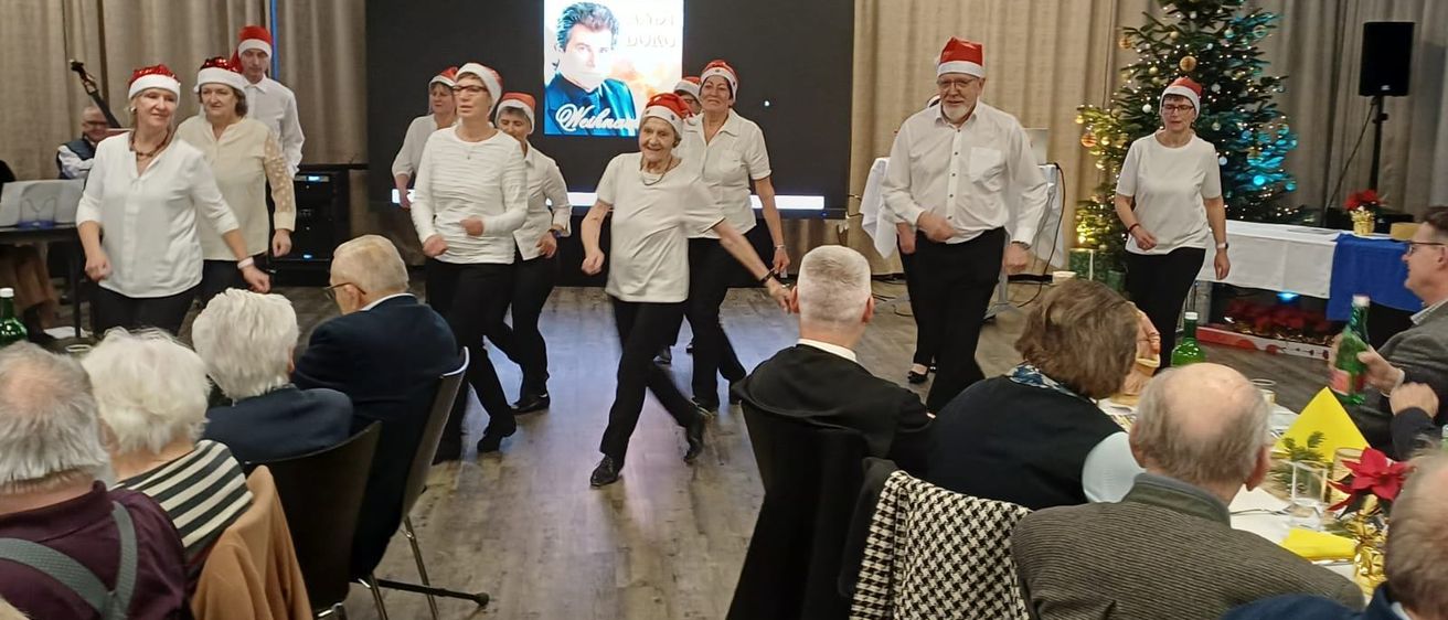 Eine Gruppe älterer Menschen in Weihnachtsmützen tanzt in einem Raum mit einem Weihnachtsbaum und einer Projektionsleinwand. Einige sitzen, andere stehen und beobachten.