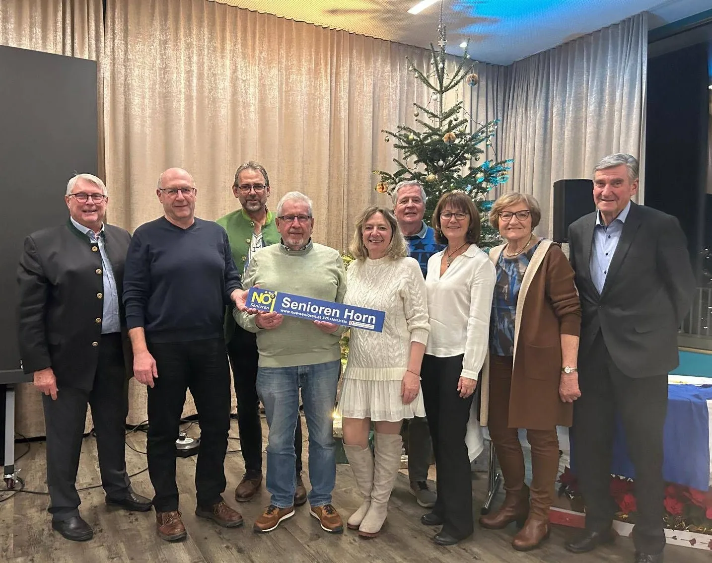 Eine Gruppe von Menschen steht gemeinsam vor einem Weihnachtsbaum. Ein Mann hält ein blaues Schild. Sie alle lächeln.