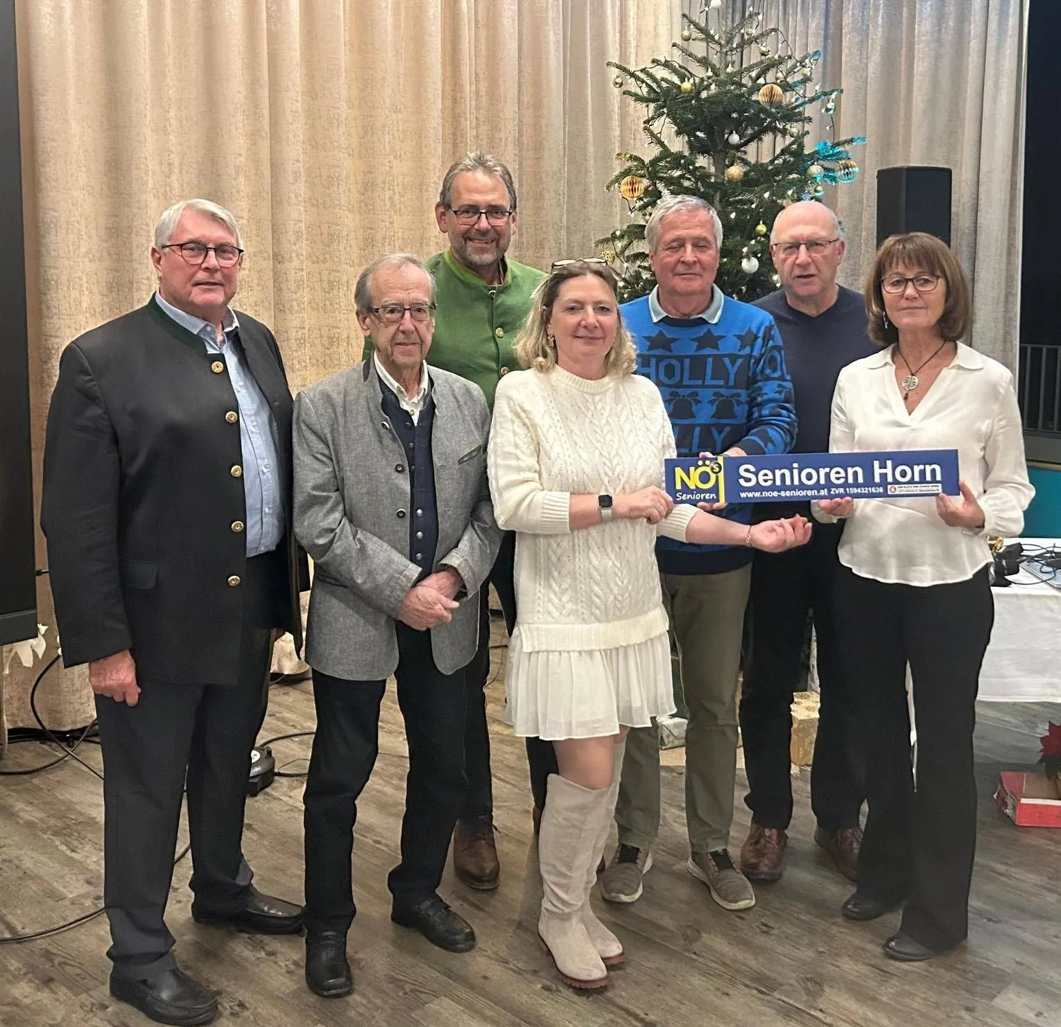 Eine Gruppe älterer Menschen, möglicherweise ein Seniorenchor, steht zusammen vor einem Weihnachtsbaum und hält ein Schild mit der Aufschrift Senioren Horn.