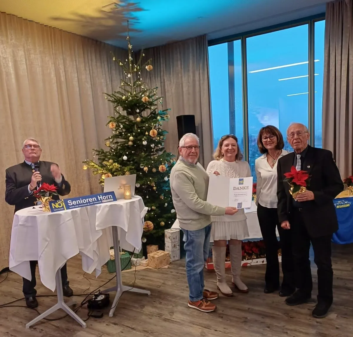 Fünf Personen stehen in einem Raum mit einem Weihnachtsbaum, einem Mann mit Mikrofon, einem Mann mit einem Zertifikat und einer Frau mit einem Blumenstrauß.
