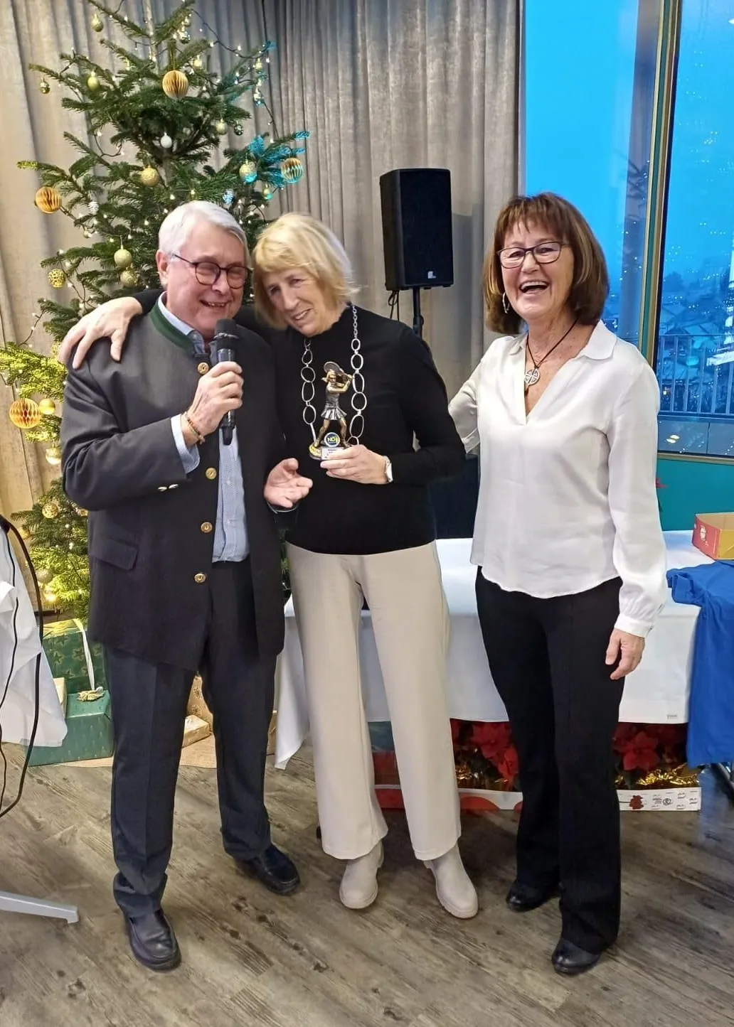 Drei Menschen stehen nebeneinander, lächeln und posieren für ein Foto. Ein Mann hält ein Mikrofon und eine Frau ein Preisschild. Dahinter steht ein Weihnachtsbaum und ein Lautsprecher auf einem Ständer.