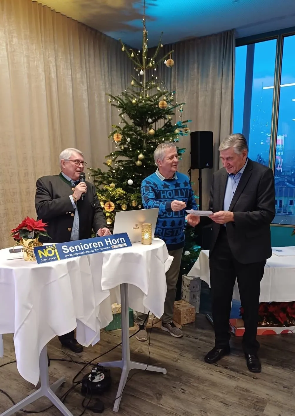 Drei Männer stehen vor einem Weihnachtsbaum, einer hält ein Mikrofon, ein anderer liest von einem Papier und der dritte lächelt. Ein Laptop und eine Kerze stehen auf dem Tisch.