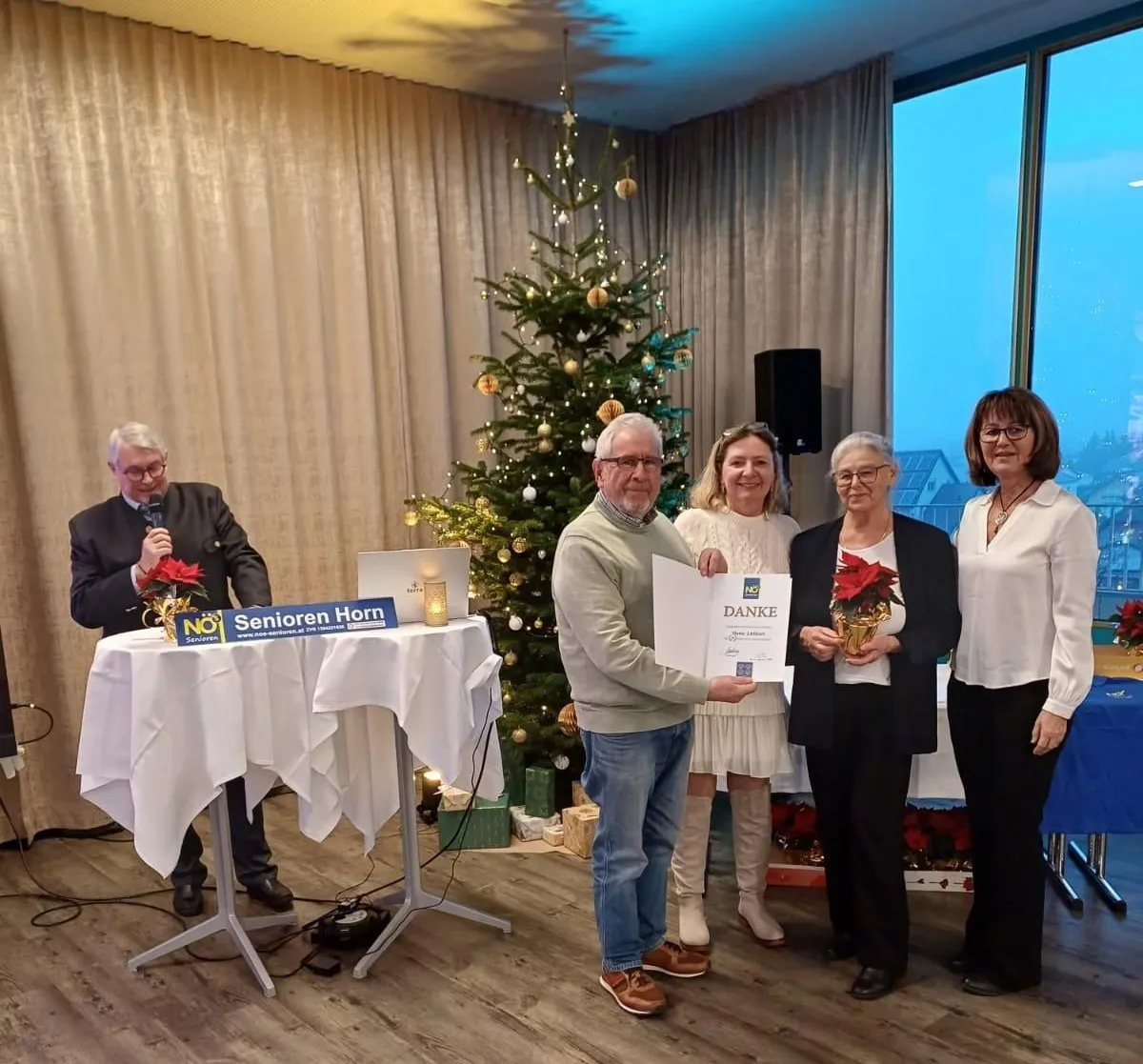 Fünf Personen stehen in einem Raum, der mit einem Weihnachtsbaum geschmückt ist, und halten ein Zertifikat und Blumen. Ein Mann mit Mikrofon steht an einem Tisch mit einem Schild. Dahinter steht ein weiterer Tisch mit einer Kerze und einem Lautsprecher.