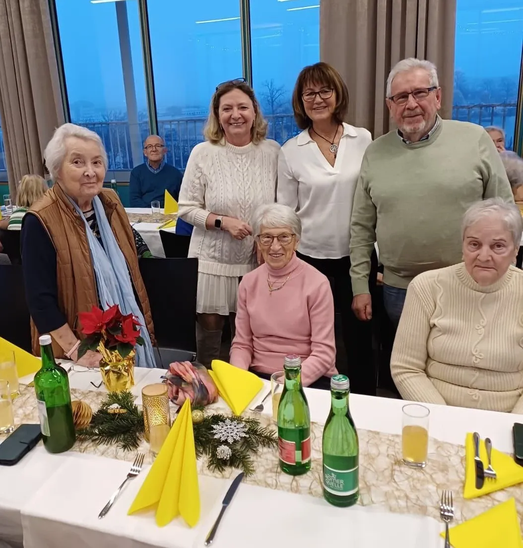 Eine Gruppe älterer Menschen sitzt an einem festlich gedeckten Tisch und lächelt in die Kamera. Sie sind von Feiertagsdekorationen und Getränken umgeben. Der Tisch ist mit Tellern, Besteck und Servietten gedeckt. Im Hintergrund sitzen und stehen Menschen in der Nähe eines großen Fensters.