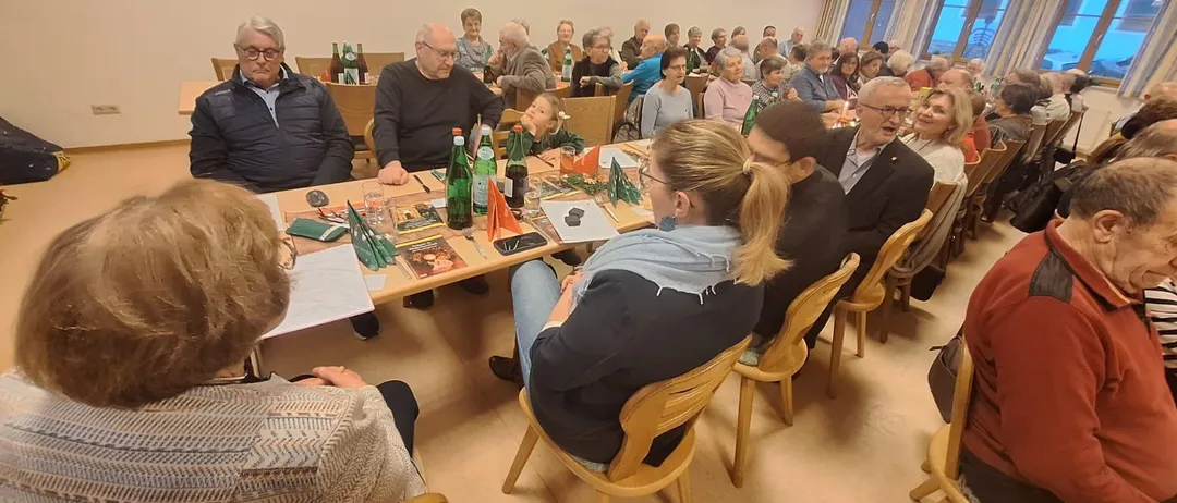 Eine Gruppe älterer Menschen sitzt um einen Tisch herum, auf dem verschiedene Gegenstände wie Flaschen, Tassen und Papiere stehen. Einige unterhalten sich. Eine Frau mit Pferdeschwanz sitzt und redet. Ein Kind sitzt vor dem Tisch. Der Raum ist gut beleuchtet.