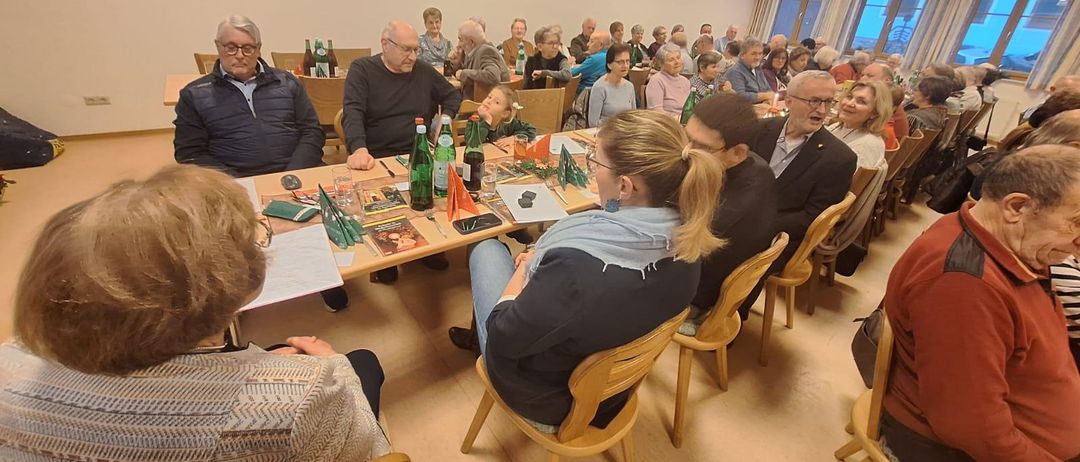 Eine Gruppe älterer Menschen sitzt um einen Tisch herum, auf dem verschiedene Gegenstände wie Flaschen, Tassen und Papiere stehen. Einige unterhalten sich. Eine Frau mit Pferdeschwanz sitzt und redet. Ein Kind sitzt vor dem Tisch. Der Raum ist gut beleuchtet.