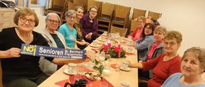 Eine Gruppe von Frauen sitzt an einem Tisch im Esszimmer und hält ein blaues Schild mit der Aufschrift 'Senioren'. Sie sind von Tellern, Tassen und Kerzen umgeben. Die Frau auf der linken Seite hält das Schild und die anderen schauen darauf.