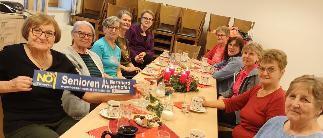 Eine Gruppe von Frauen sitzt an einem Tisch im Esszimmer und hält ein blaues Schild mit der Aufschrift 'Senioren'. Sie sind von Tellern, Tassen und Kerzen umgeben. Die Frau auf der linken Seite hält das Schild und die anderen schauen darauf.
