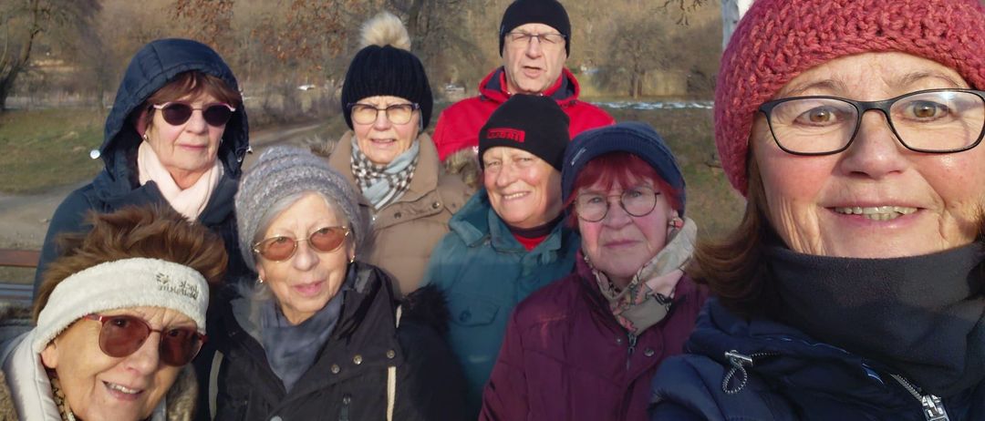 Eine Gruppe älterer Personen in Winterkleidung steht in einem Park mit einer malerischen Aussicht auf Hügel und Bäume unter einem blauen Himmel mit einigen Wolken.