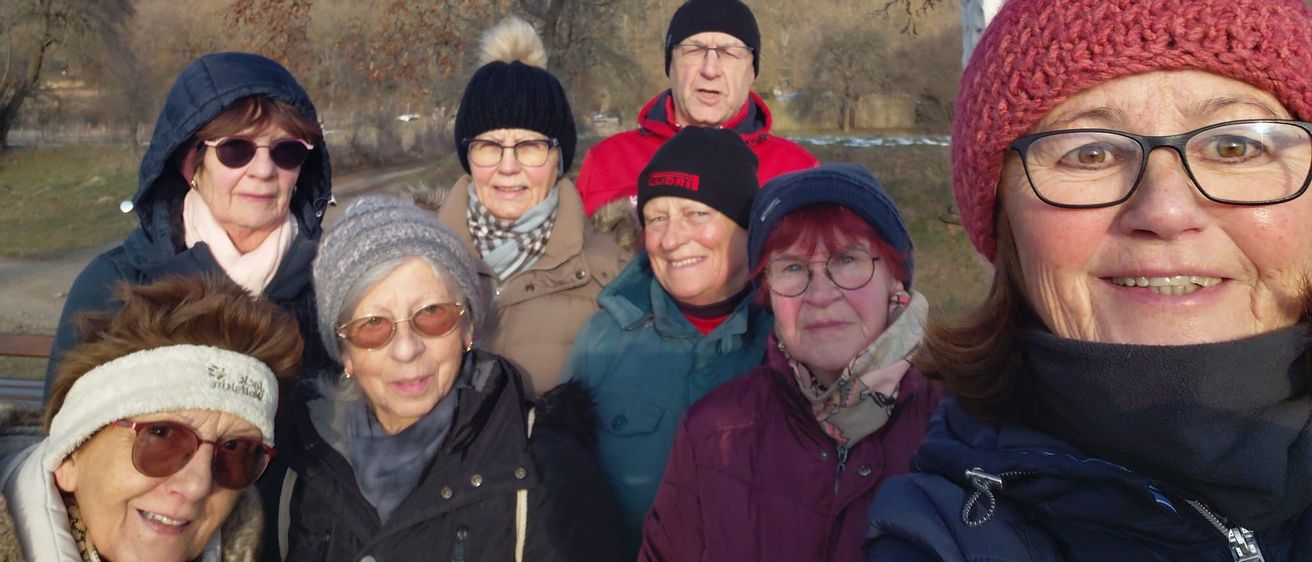 Eine Gruppe älterer Personen in Winterkleidung steht in einem Park mit einer malerischen Aussicht auf Hügel und Bäume unter einem blauen Himmel mit einigen Wolken.