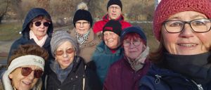 Eine Gruppe älterer Personen in Winterkleidung steht in einem Park mit einer malerischen Aussicht auf Hügel und Bäume unter einem blauen Himmel mit einigen Wolken.