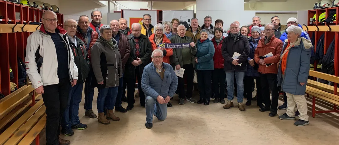 Eine Gruppe von Menschen steht in einem Raum zusammen. Einige halten Papiere und lächeln für ein Foto. Ein Banner hinter ihnen lautet 'Senioren Horn'.