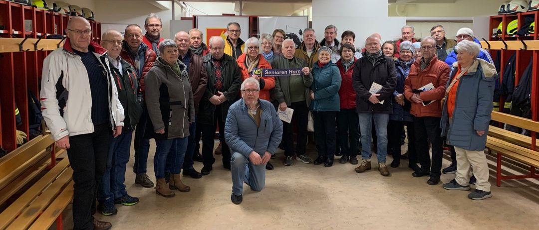 Eine Gruppe von Menschen steht in einem Raum zusammen. Einige halten Papiere und lächeln für ein Foto. Ein Banner hinter ihnen lautet 'Senioren Horn'.