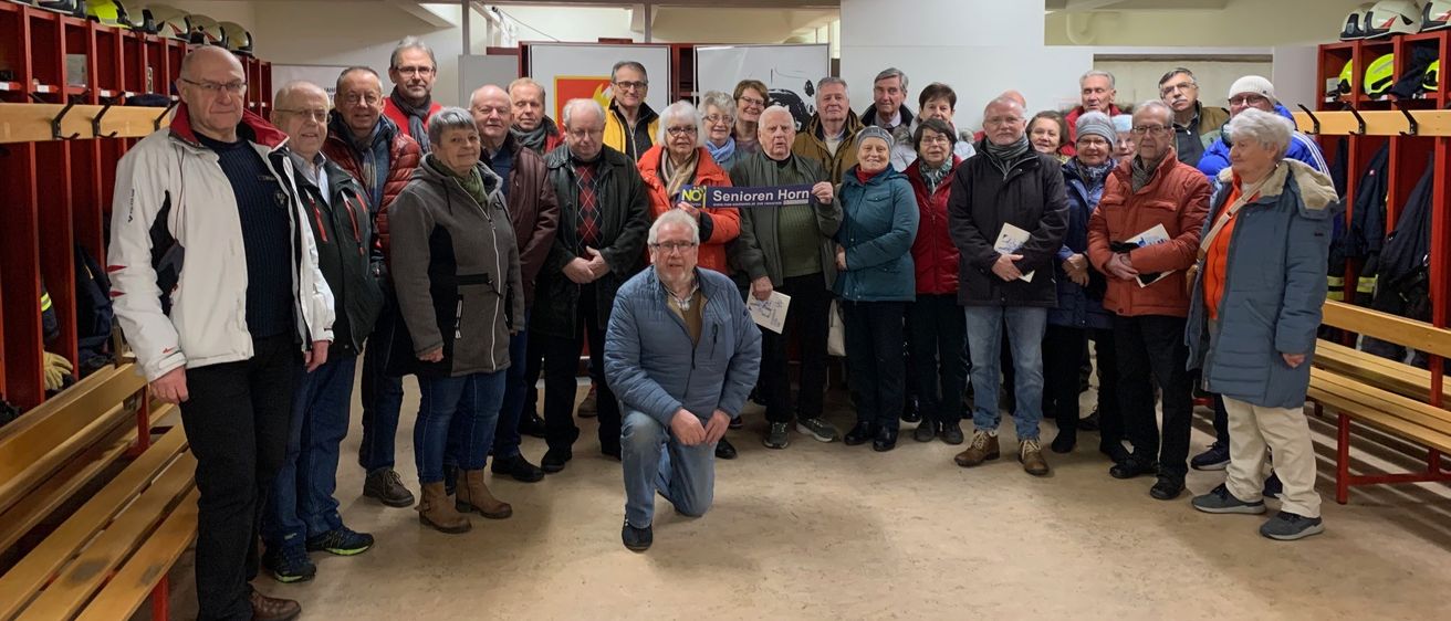 Eine Gruppe von Menschen steht in einem Raum zusammen. Einige halten Papiere und lächeln für ein Foto. Ein Banner hinter ihnen lautet 'Senioren Horn'.