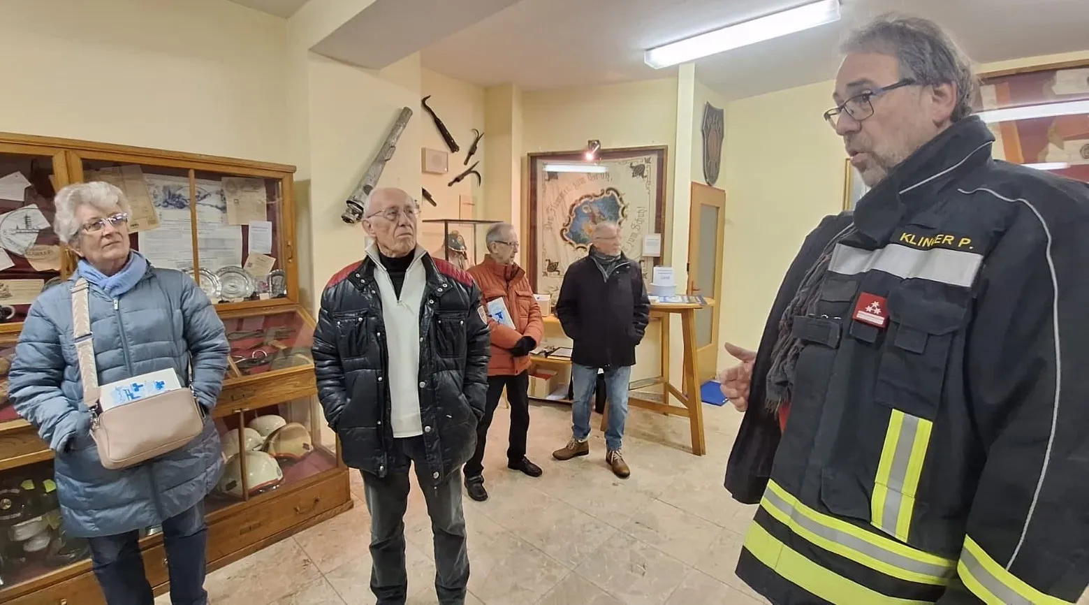 Mehrere ältere Männer stehen in einem Raum, wahrscheinlich einem Museum, mit Ausstellungen im Hintergrund. Ein Mann trägt eine schwarze Lederjacke.