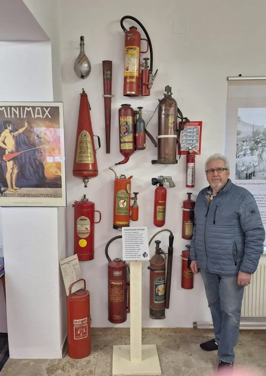 Ein Mann steht vor einer Wand mit verschiedenen roten Feuerlöschern in unterschiedlichen Formen und Größen. Ein Plakat mit einem Mann in einem roten Umhang befindet sich auf der linken Seite.