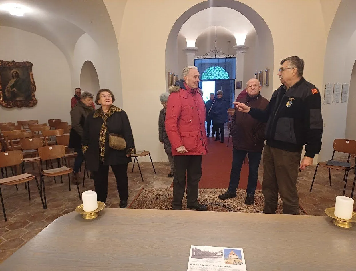 Eine Gruppe von Menschen ist in einem Raum, ein Mann in einem roten Mantel spricht. Ein Mann mit Brille zeigt. Eine Kerze steht auf einem Tisch.