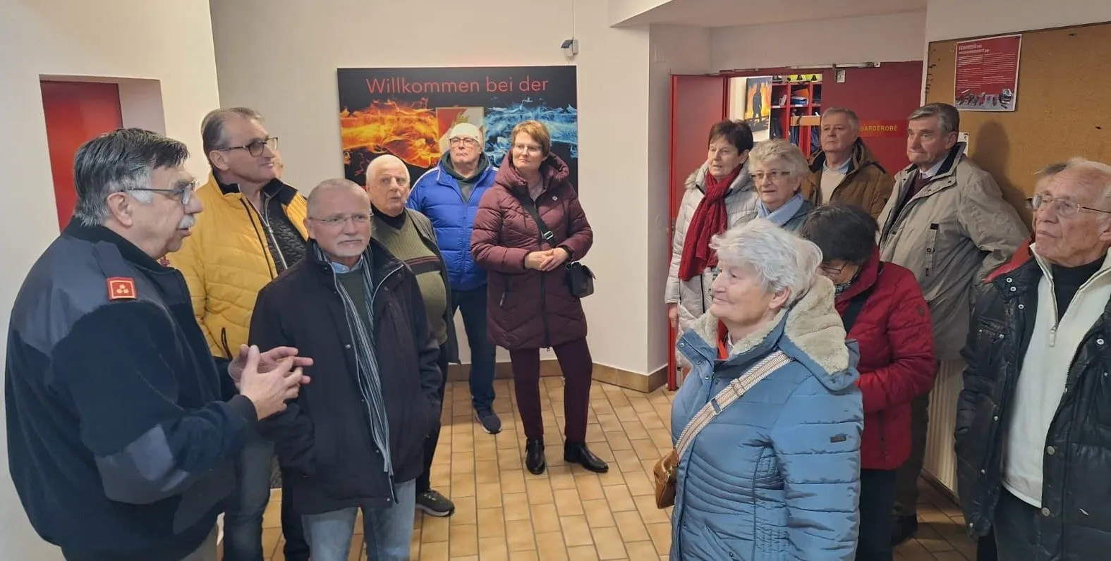 Eine Gruppe von Menschen steht in einem Raum mit einem Feuer- und Wasser-thematischen Plakat. Sie tragen Winterkleidung und scheinen sich zu unterhalten.