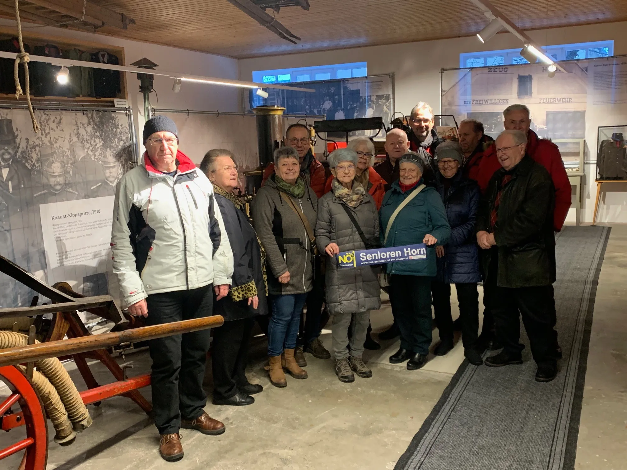 Eine Gruppe älterer Menschen steht lächelnd für ein Foto in einem Raum. Ein Schild sagt 'NEIN! Senioren Horn'. Der Raum hat eine Holzdecke, Poster und Lampen.