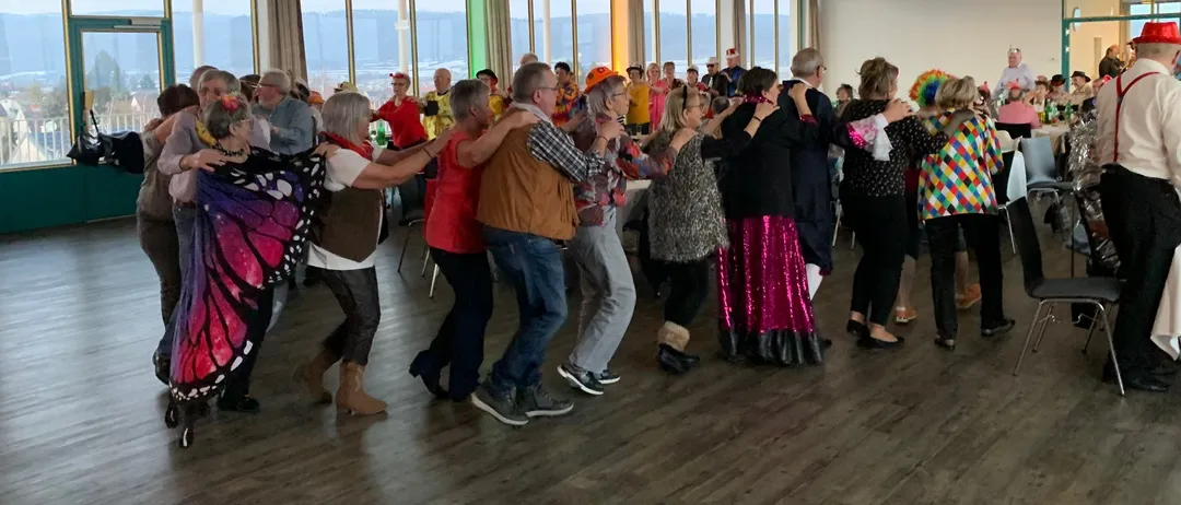 Eine Gruppe von Menschen in verschiedenen Kostümen tanzt gemeinsam in einem großen Raum mit Holzböden und großen Fenstern.